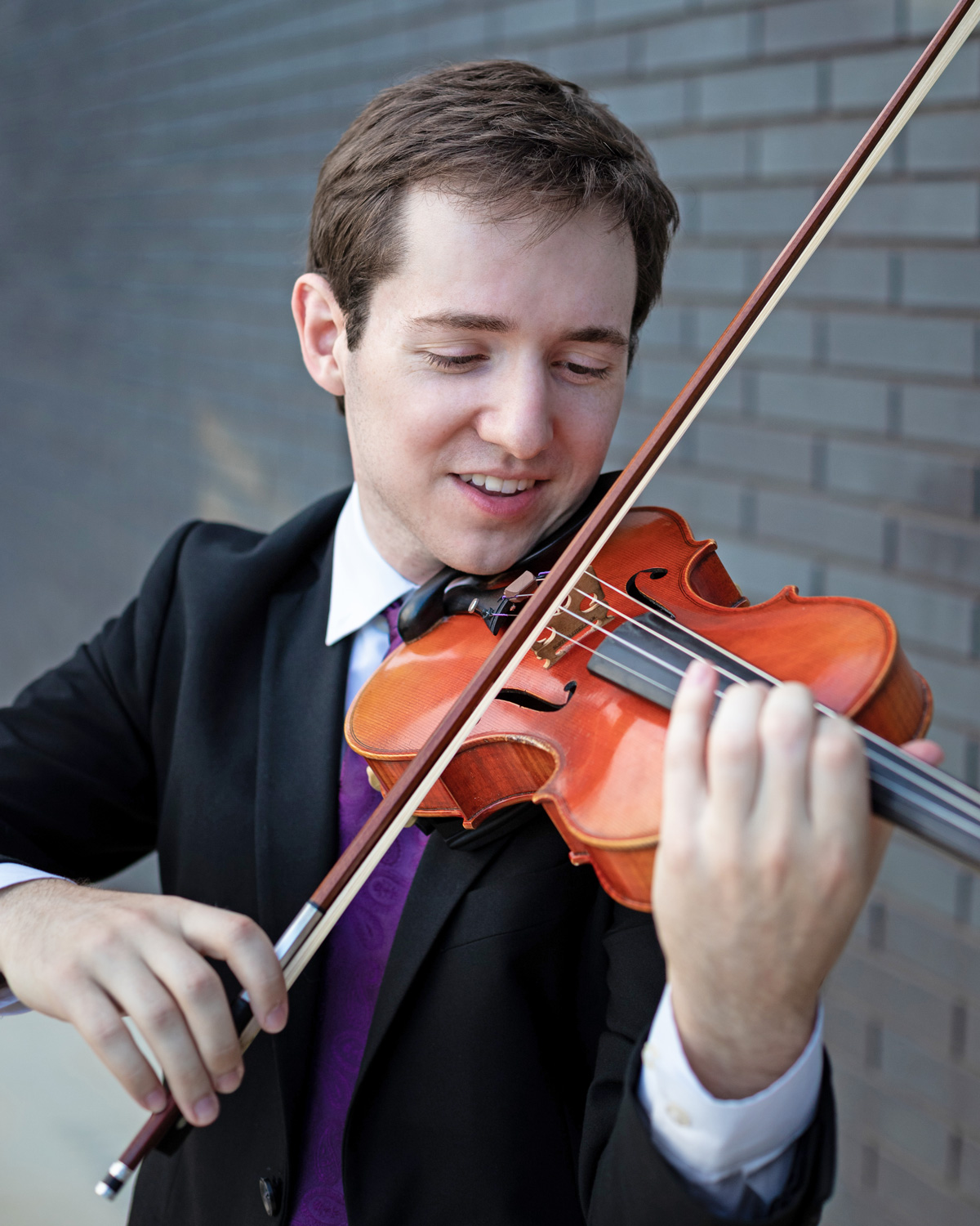 a young smiling man plays violin. he is wearing a suit and a purple tie.