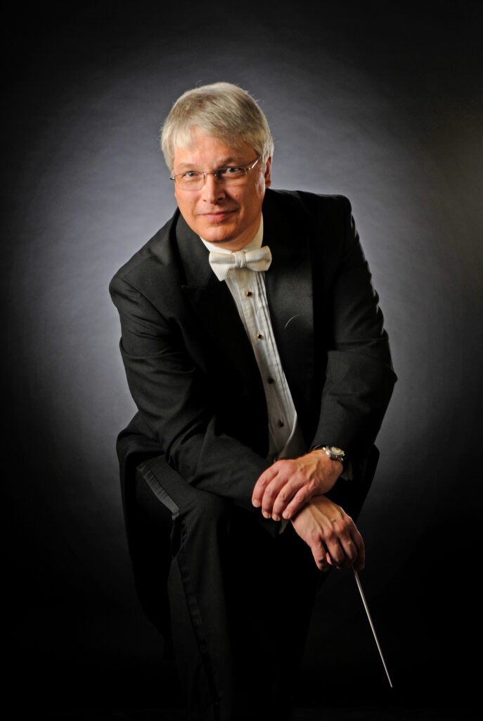 a man with a tuxedo, gray hair and a white bow tie sits with his hands crossed on his leg