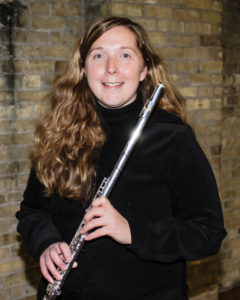 A young caucasian woman with very long blonde hair is dressed in black. She smiles and holds a flute.