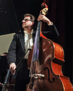 A middle-aged caucasian man with glasses and dark curly hair wears a tuxedo and plays a red upright bass.
