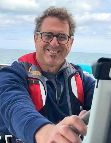 A middle-aged caucasian man with glasses and curly graying hair steers a boat 
