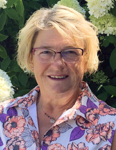 A middle-aged caucasian woman with glasses and medium length blonde hair in front of a hydrangea.