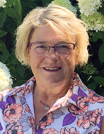 a Caucasian woman with glasses and medium length blonde hair in front of a hydrangea
