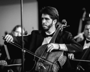 A black and white photo of a caucasian adult man with dark hair and a beard playing cello. He wears a tuxedo.