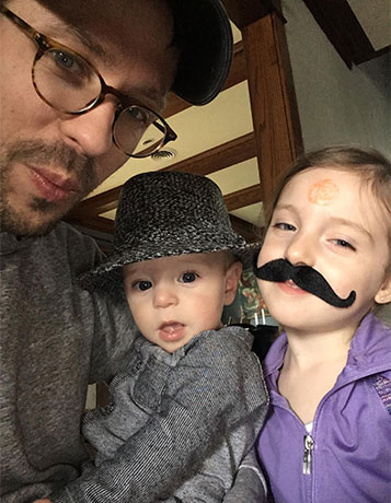 selfie of a young Caucasian man with a mustache and glasses with a baby boy in an adult's hat and a young girl with a fake handlebar mustache