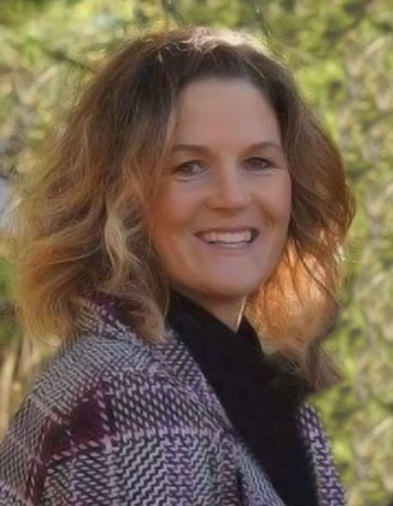 A smiling caucasian woman with a plaid coat and brown shoulder length hair in front of trees.