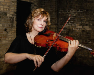 A middle-aged caucasian woman with blonde hair wears a black dress and plays a viola.