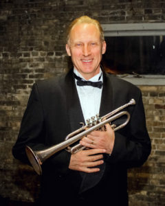 A middle-aged caucasian man with light hair dressed in a tuxedo. He smiles and holds a trumpet.