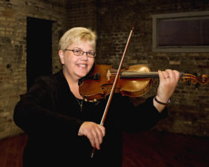 A middle-aged caucasian woman with short blonde hair smiles and plays a violin. She is dressed in black.
