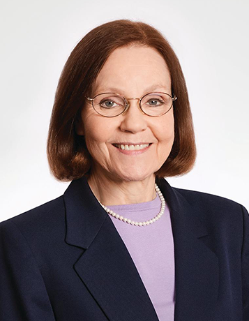 A smiling mature caucasian woman with medium length brown hair and glasses wears a dark jacket and lavender blouse and pearls.