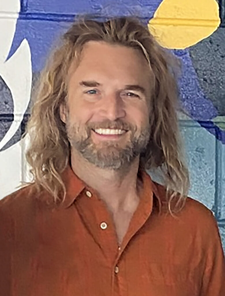 A man with long blonde hair and beard wearing an orange shirt stands in front of a colorfully painted block wall.