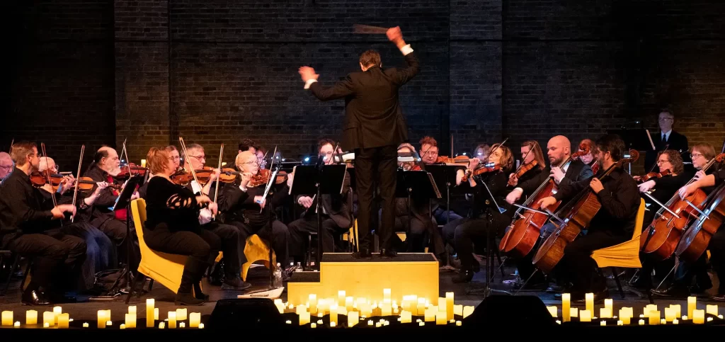 An orchestra of stringed instrument players seated with a male conductor standing with his back to the camera. Candles line the stage.