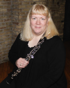 A middle-aged caucasian woman with long blonde hair is dressed in black. She smiles and holds an oboe.