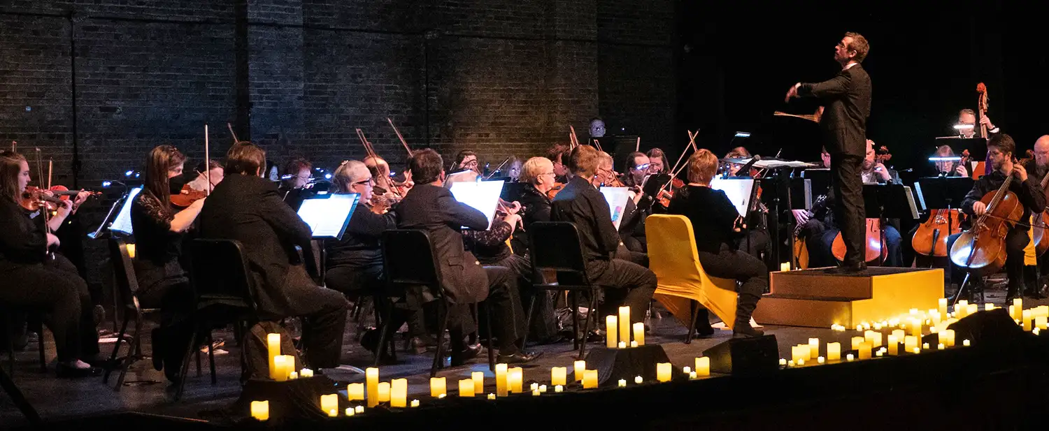 An orchestra on a dark stage with a male conductor and glowing candles lining the stage.